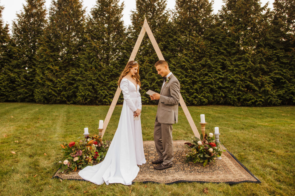 Summer 2022 bride and groom reading wedding vows in front of a wooden triangle arch. 
